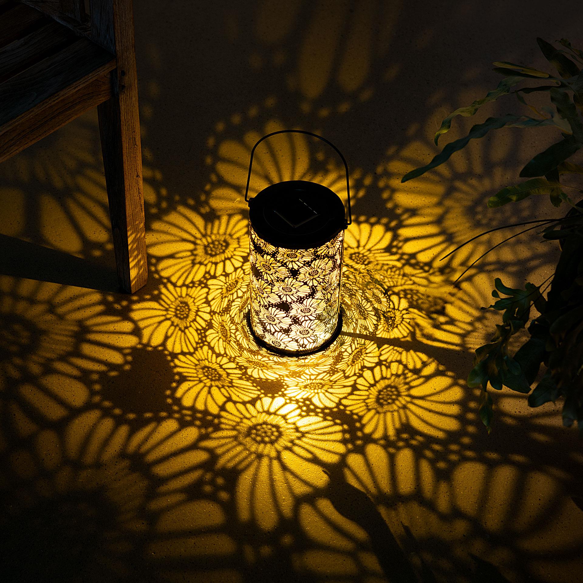 Eine zylindrische Solarlaterne mit ausgestanztem Gerbera-Blumenmuster und einem dünnen Metallbügel zum Aufhängen