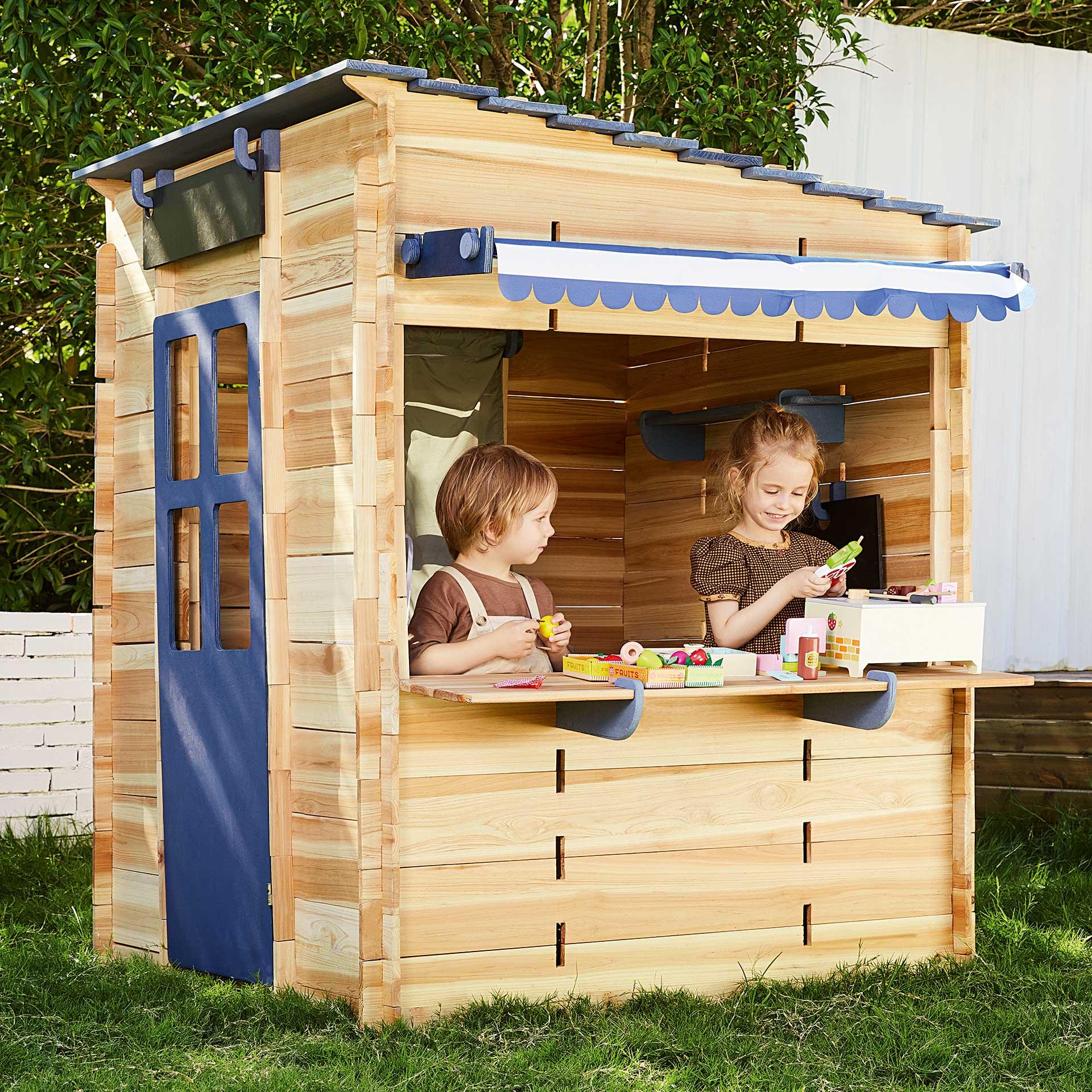 Ein Gartenspielhaus aus Holz mit blauen Teilen, Tür, Theke, Sonnenschutz und Fenster mit Fenstersims steht im Garten