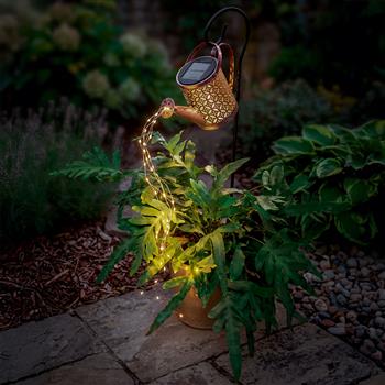 Eine bronzefarbene Solarlicht Giesskanne an einem schwarzen Metallhaken, aus deren Ausguss leuchtende LED-Drahtlichter wie Wasserstrahlen fliessen